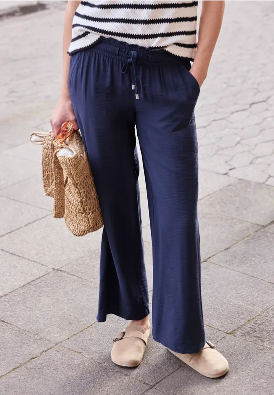 Persoon met wijde marineblauwe broek, gestreept shirt, rieten tas en beige espadrilles op het trottoir.