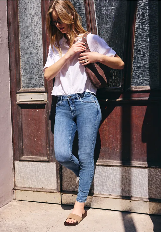 Femme adossée à une porte, en t-shirt blanc, jean et sac à dos, regard vers le bas.