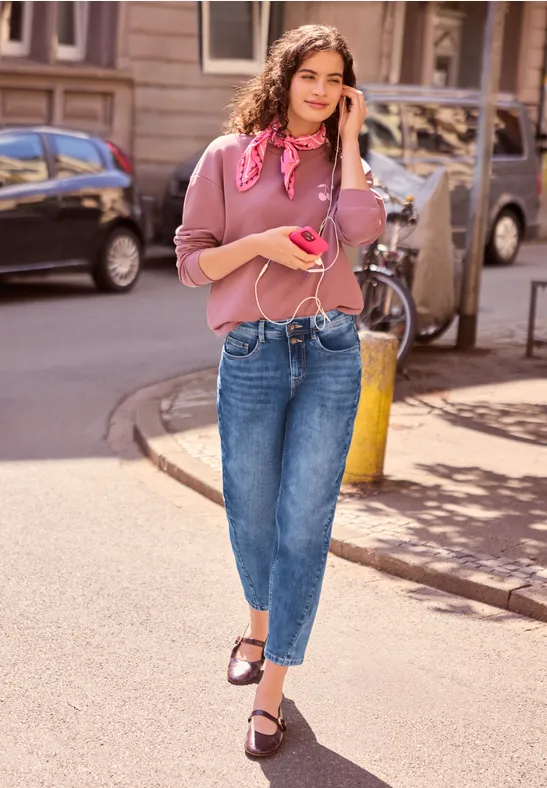 Junge Frau in rosa Oberteil und Jeans mit rosa Handy telefoniert fröhlich beim Spaziergang.