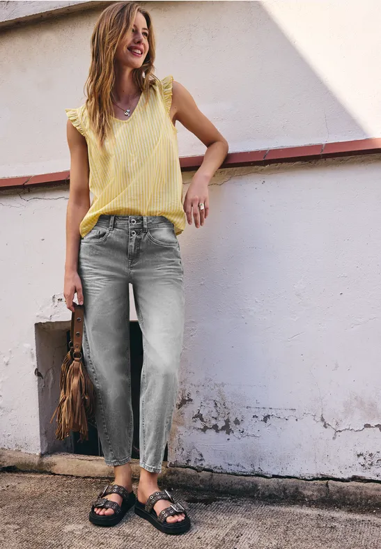 Femme en haut jaune, jean gris et sandales noires, appuyée nonchalamment contre un mur blanc.
