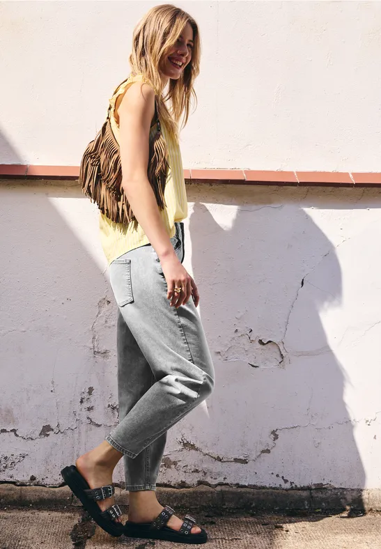 Une jeune femme en top jaune, pantalon gris et sandales noires, appuyée décontractée contre un mur blanc.