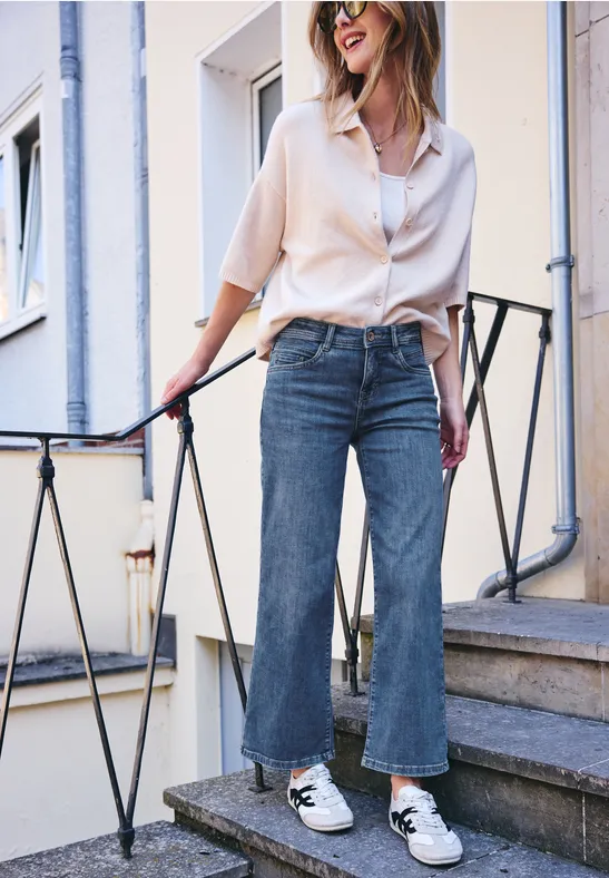 Une femme posant sur un escalier porte une chemise blanche ample, un jean à jambes larges et des baskets blanches.