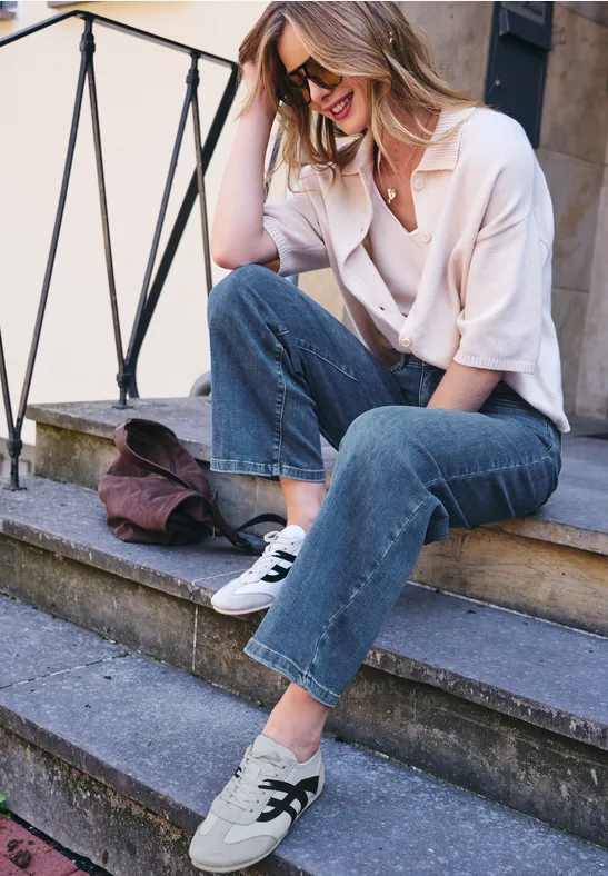 Femme assise sur des marches, blouse claire, jean et baskets blanches à étoile, souriante et détendue.