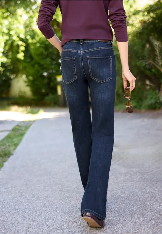 Person in dunkelblauen Jeans und Pullover geht über einen Gartenweg, hält eine Sonnenbrille