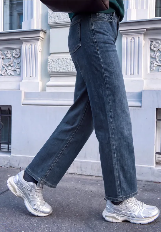Beine in weiten Jeans und silbernen Sneakern beim Gehen vor einer Hausfassade.