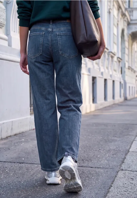 Person in blauen Jeans, Turnschuhen und Tasche läuft auf Gehweg entlang eleganter Häuser.