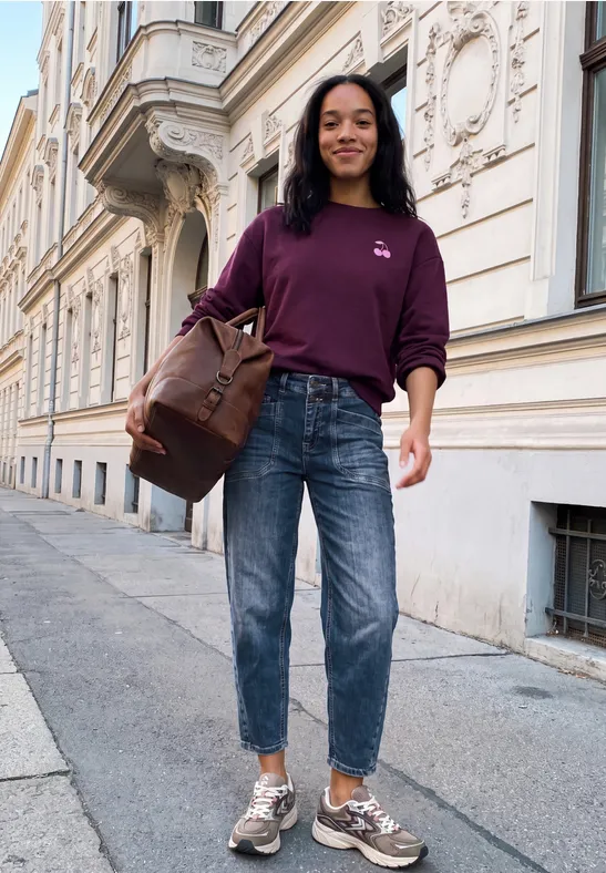 Kobieta w fioletowym swetrze, jeansach i sneakersach niesie dużą torbę, idąc uśmiechnięta przed zabytkową kamienicą.
