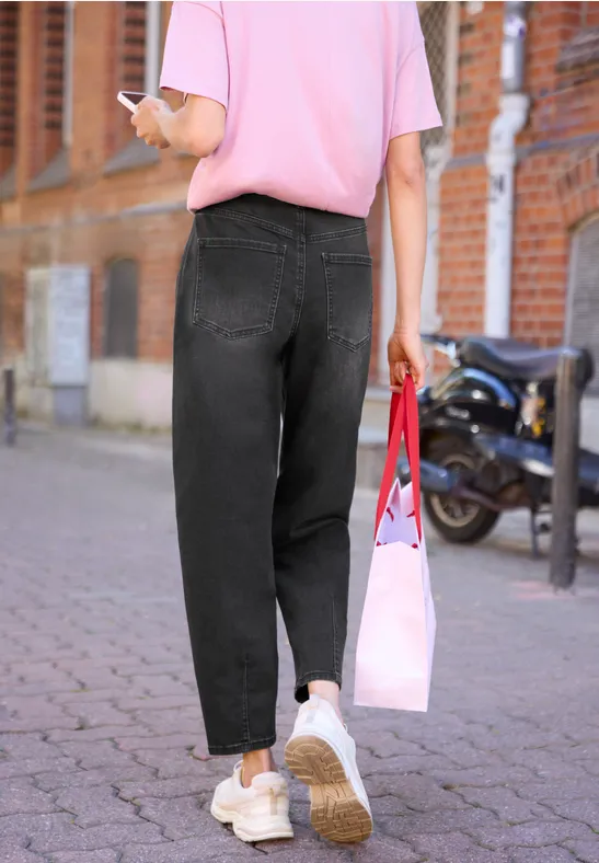 Person geht auf gepflastertem Gehweg, trägt rosa T-Shirt, schwarze Jeans, weiße Sneaker, hält Handy und rosa Tasche.