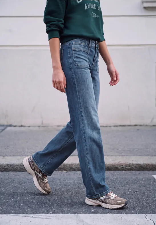 Person in blauer Jeans, grünem Pullover und Sneakers geht auf Gehweg vor heller Wand.