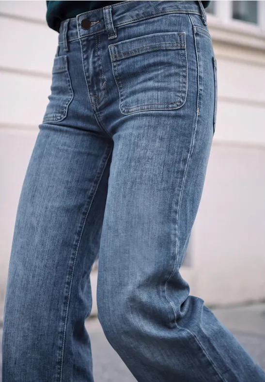 Seitliche Nahaufnahme einer Person in lockeren, blauen Jeans mit aufgesetzten Taschen vor hellem Hintergrund.