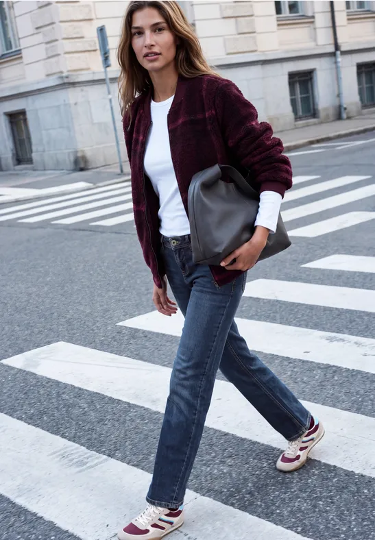 Eine junge Frau in Jeans, weißem Shirt und bordeauxrotem Cardigan überquert eine städtische Straße mit Handtasche.
