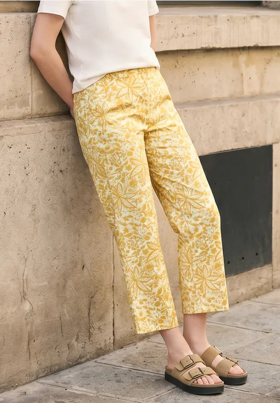 Een vrouw leunt tegen een muur, draagt een wit shirt, gele broek met bloemmotief en groene sandalen.
