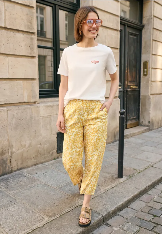 Persoon in wit T-shirt en gele bloemenbroek staat op de stoep voor een stenen gevel.