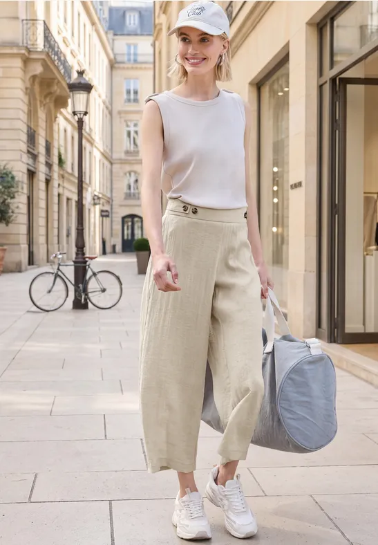 Vrouw in wit topje, wijde beige broek en sneakers met reiskoffer loopt door een stijlvolle straat.