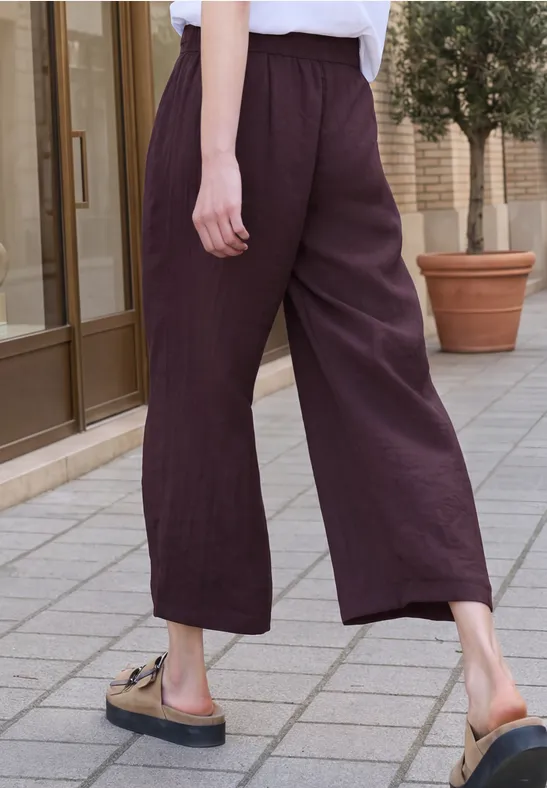 Eine Person trägt weite, dunkelviolette Leinenhose und Sandalen und geht auf einem gepflasterten Bürgersteig.