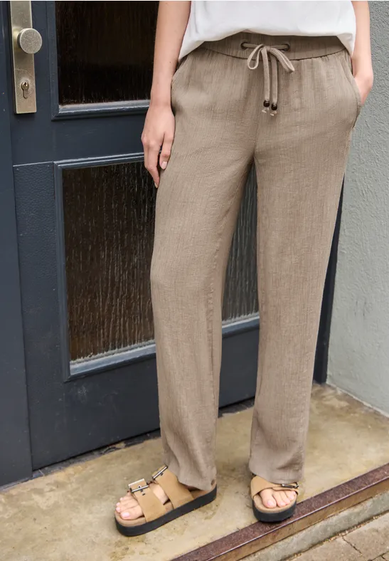 Helle, lockere Leinenhose mit Tunnelzug und Taschen, kombiniert mit beigen Sandaletten auf Schrittstufe vor Tür.