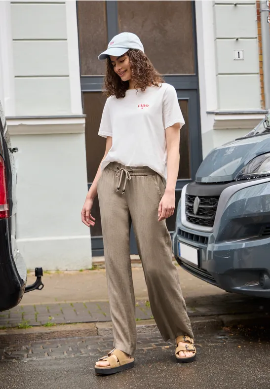 Junge Frau mit weißer Kappe, T-Shirt und weiten Stoffhosen steht lässig zwischen Autos auf dem Gehweg.
