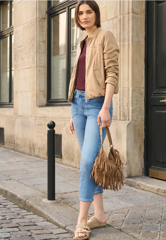 Vrouw in casual outfit met beige jasje, bordeaux top, blauwe jeans en franjes tas op kasseienstraat.