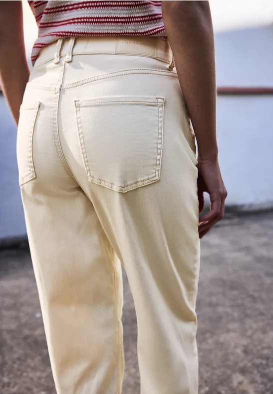 Vrouw van achteren in crème broek met zakken en gestreept topje, geleund tegen reling.