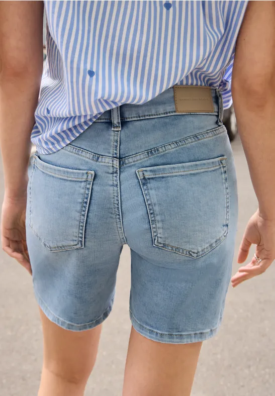 Person von hinten in hellblauen Jeans-Shorts und gestreiftem Hemd auf hellem Untergrund.