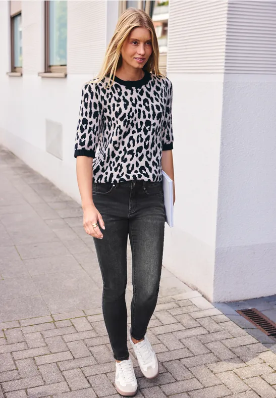 Junge Frau in Leopardenpullover, dunklen Skinny-Jeans und weißen Sneakern geht mit einem Ordner die Straße entlang.