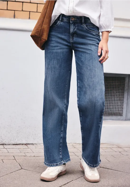Eine Person trägt weite blaue Jeans, weiße Schuhe, weiße Bluse und trägt eine braune Tasche.