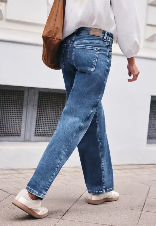 Eine Person in blauen Jeans und hellen Schuhen mit brauner Umhängetasche geht an einer Häuserwand entlang.