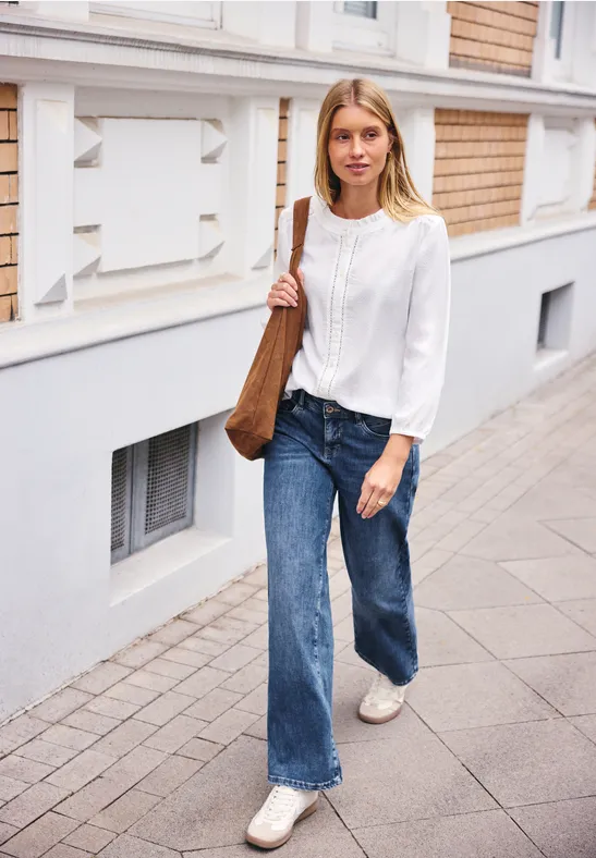 Eine junge Frau in weißer Bluse, blauen Jeans und brauner Tasche geht auf dem Gehweg an einem Gebäude vorbei.