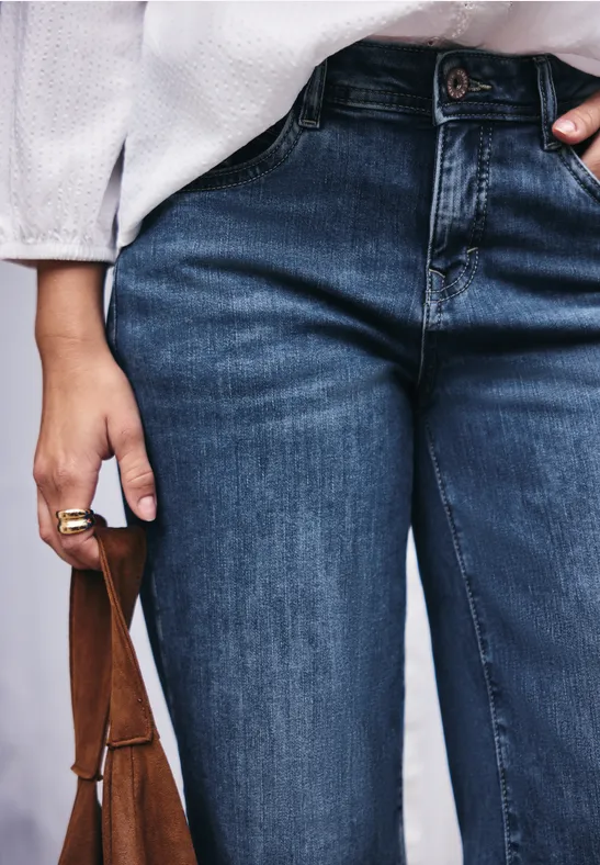 Person trägt dunkelblaue Jeans, weiße Bluse und hält eine braune Ledertasche in der Hand.
