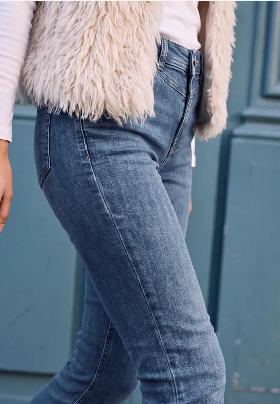 Eng anliegende blaue Jeans und weiße flauschige Weste vor einer türkisfarbenen Tür.