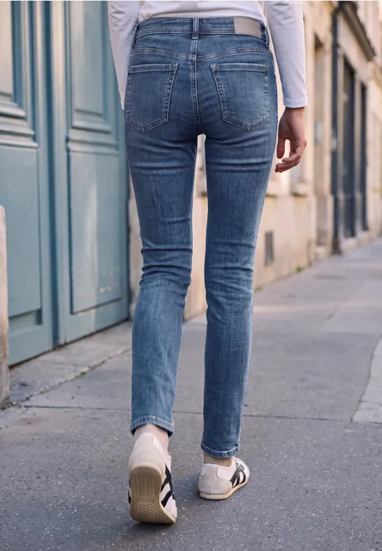 Eine Person von hinten in blauen Skinny-Jeans und weißen Sneakern, die auf einem Gehweg in der Stadt läuft.