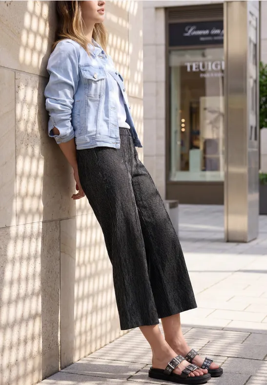 Frau in heller Jeansjacke und weiten schwarzen Hosen lehnt vor Schaufenster und gemusterten Säulen.