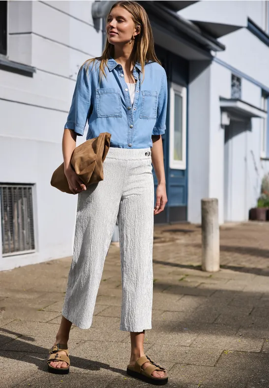 Frau in Jeanshemd, weißen Leinenhosen und Keilabsatz-Sandalen geht mit Strohhut spazieren.