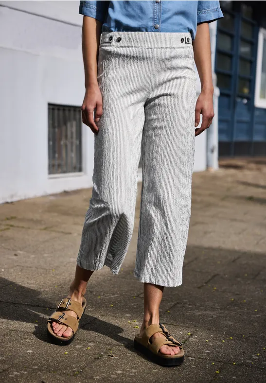 Beine einer Frau in gestreiften Leinen-Culottes und Sandalen, stehend auf einer Straße vor Hauswand.