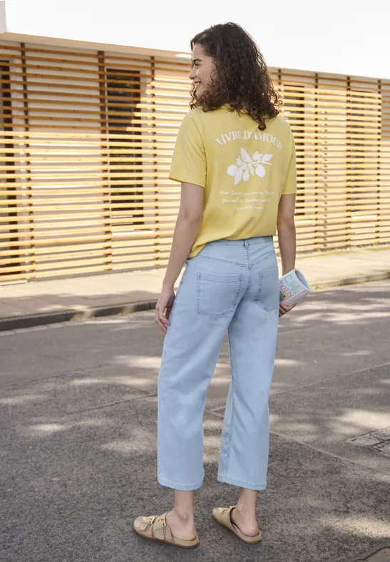 Persoon in geel t-shirt met bloemenprint, lichtblauwe jeans en sandalen loopt langs een houten schutting.