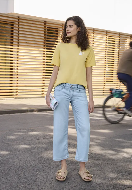 Vrouw in geel T-shirt en lichtblauwe jeans met telefoon, lopend voor een houten schutting, fietser achter haar.