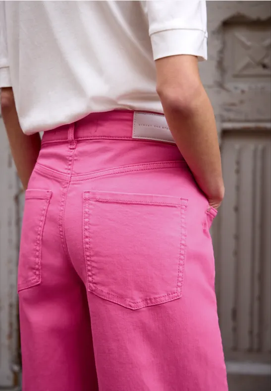 Nahaufnahme einer Person von hinten, trägt ein weißes Top und knallpinke Hose, steht vor einer grauen Metalltür.