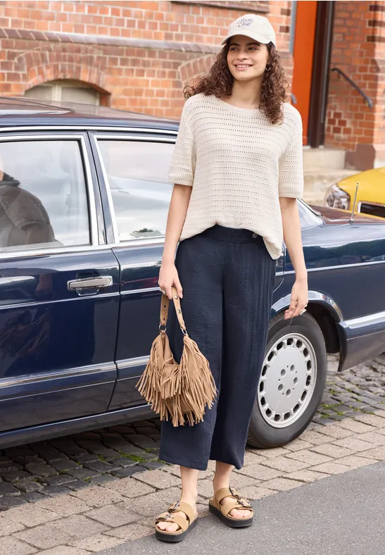Eine Frau mit Fransen-Handtasche und Sandalen steht vor einem dunkelblauen Auto auf dem Gehweg.