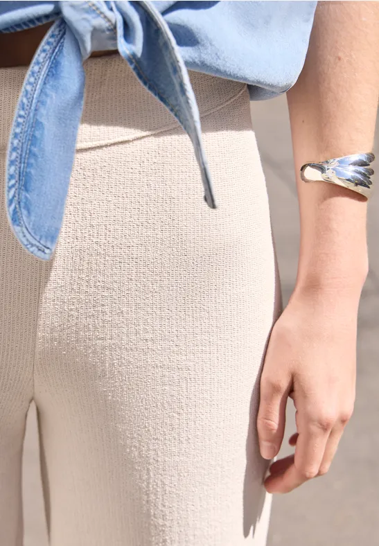 Seitliche Nahaufnahme einer Person in beige Hosen und blauem, vorne geknotetem Oberteil mit Armreif.