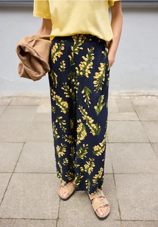 Vrouw draagt wijde zwarte broek met geel bloemenpatroon, gecombineerd met een geel shirt en sandalen.