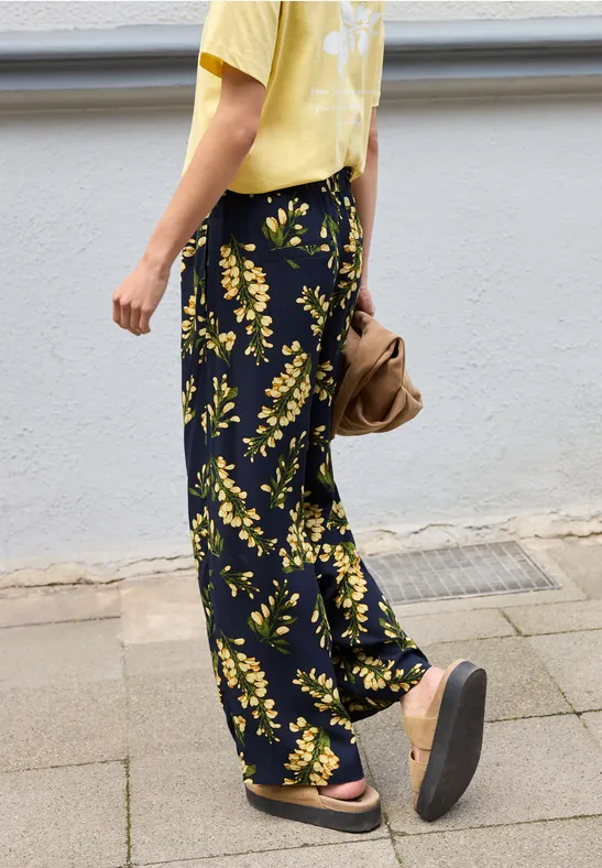 Persoon loopt op trottoir, geel t-shirt, zwarte wijde broek met gele bloemen en platformsandalen.