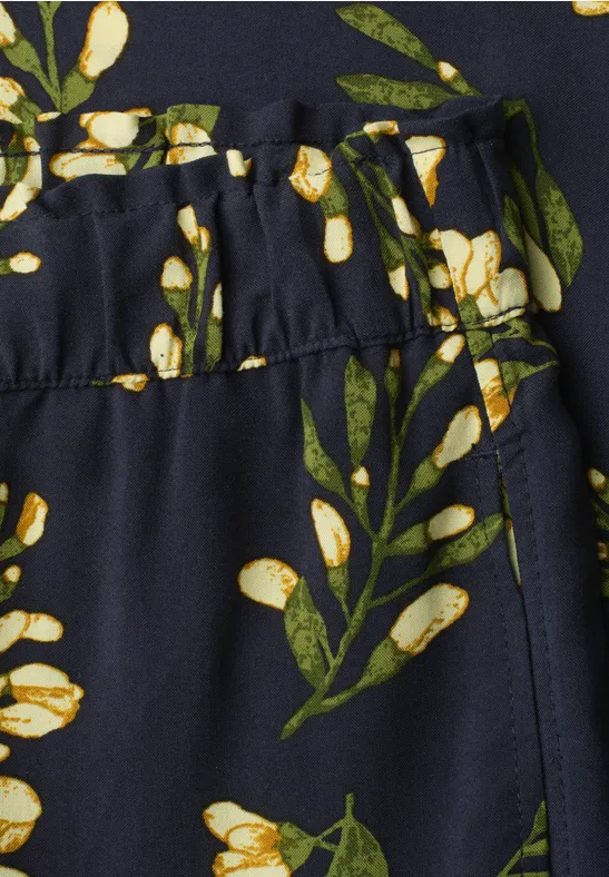Detail van een donkere stof met groene olijftakken en gele vruchten, gerimpelde tailleband.