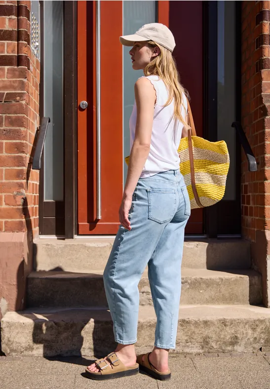 Eine Frau mit weißem Top, Cap, Jeans und Sandalen trägt eine gelb-weiße Tasche und steht vor einer Haustür.