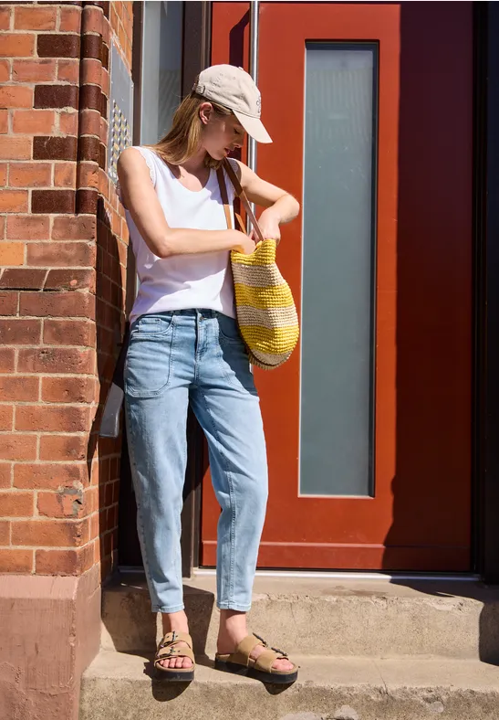 Junge Frau in Jeans, weißem Tanktop und Cap steht an einer Backsteinwand und blickt auf ihre Uhr, Tasche in der Hand.