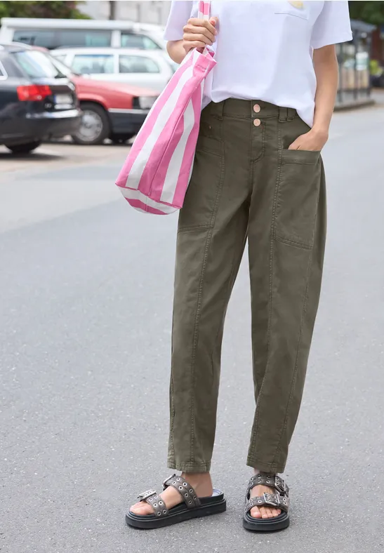 Person steht auf der Straße, trägt weite olivgrüne Hosen, weißes T-Shirt, schwarze Sandalen und eine rosa-weiße Tasche.