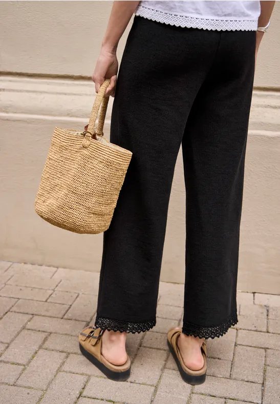 Frau trägt weit geschnittene schwarze Caprihose mit Spitzenbesatz, Basttasche und Sandalen auf Bürgersteig.