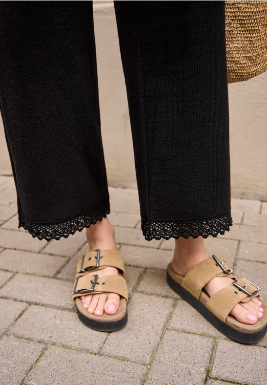 Ein Paar Füße in goldenen Sandalen mit zwei Schnallen steht auf gepflastertem Boden.