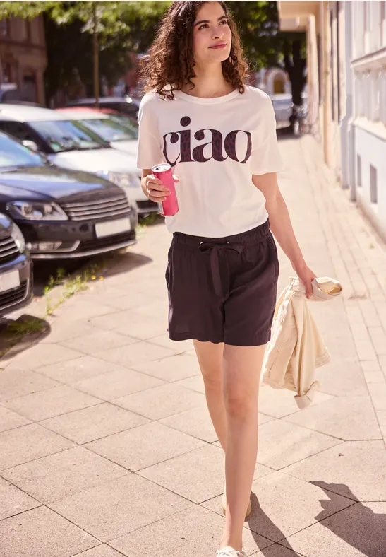Een vrouw met krullen in een wit 'ciao'-shirt loopt zonnig over straat, met drankje en jas in haar hand.