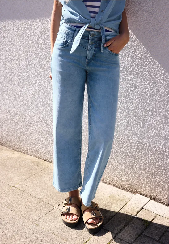 Une personne en jean large bleu et sandales noires se tient sur le trottoir devant un mur clair.