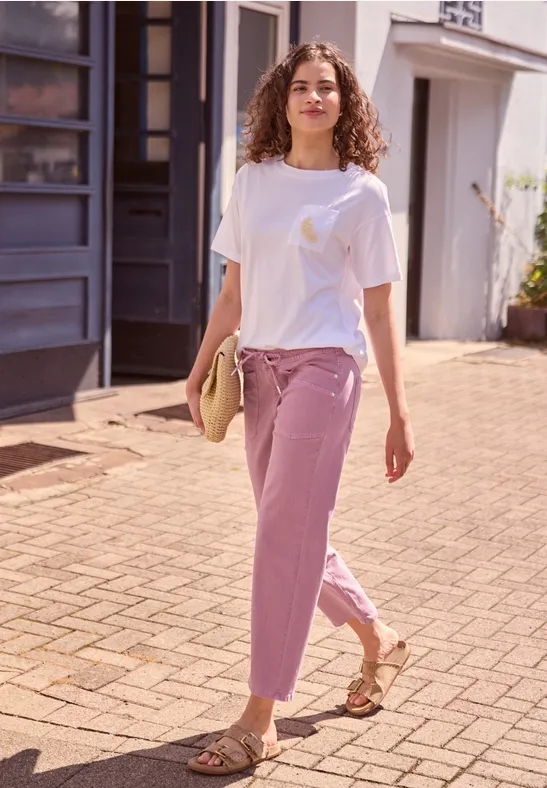 Eine junge Frau in weißem T-Shirt und lila Hose schlendert sommerlich durch die sonnige Gasse.